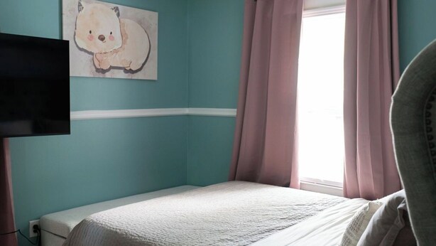 Ein Schlafzimmer mit türkisfarbenen Wänden, einem Bett mit weißer Bettdecke, einem rosa Vorhang und einem Bärenbild.