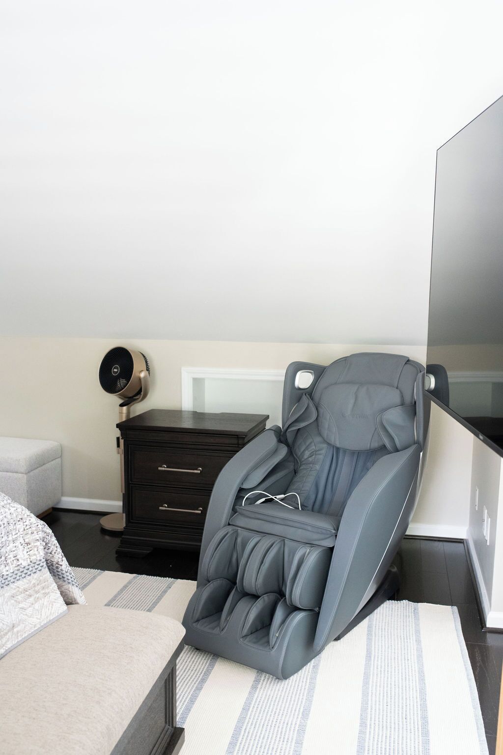 Un fauteuil de massage dans une chambre à coucher, avec un lit, une commode et un ventilateur fixé au mur.