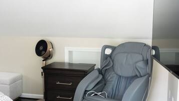 A massage chair in a bedroom with a bed, dresser, and a wall-mounted fan.