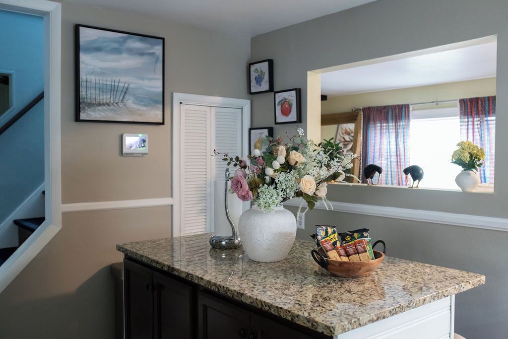 A kitchen with granite countertops, a vase with flowers, a fruit bowl, and framed pictures on the wall.