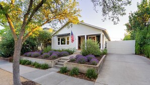 A single-story house with a front porch, a flag, and a landscaped garden with purple flowers. - Lavender Porch - Designer Bungalow with Fire Pit Near Downtown Paso (Paso Robles)