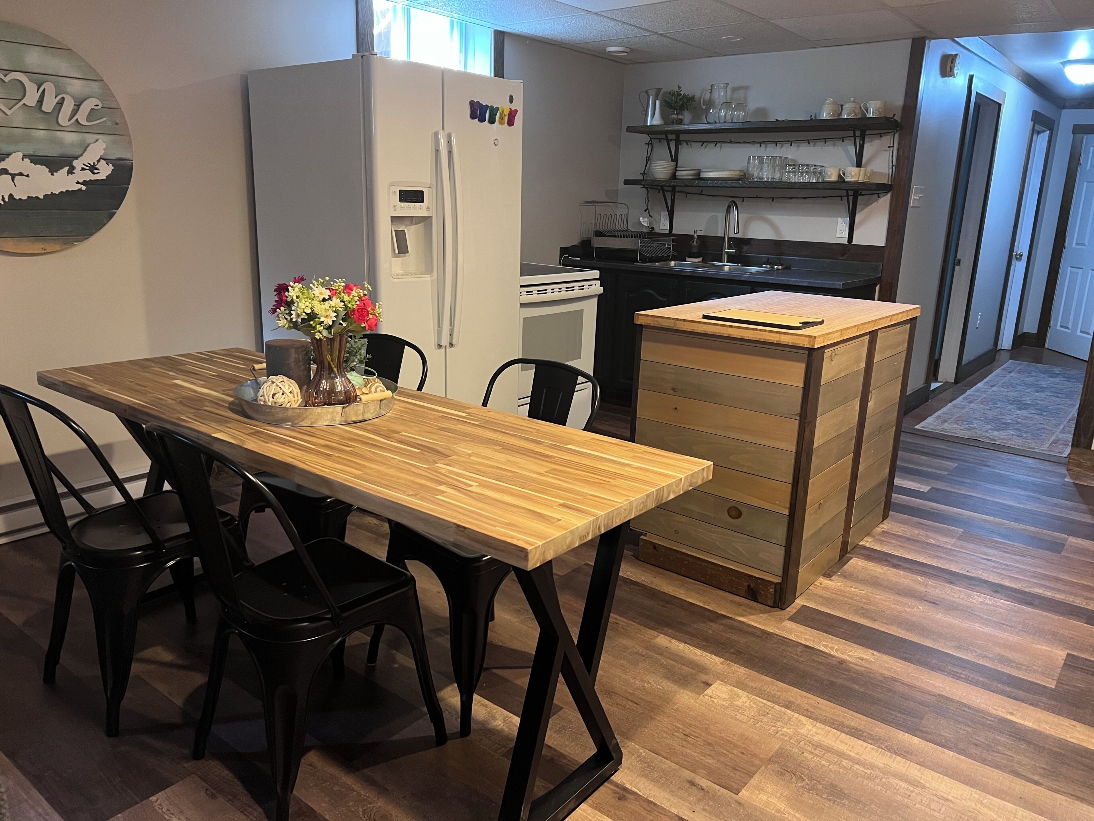 A kitchen with a refrigerator, a wooden dining table with chairs, a small island with drawers, and a shelf with dishes.