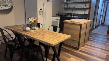 A kitchen with a refrigerator, a wooden dining table with chairs, a small island with drawers, and a shelf with dishes.