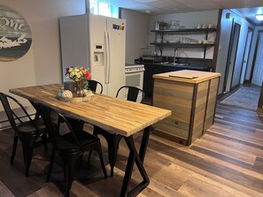 A kitchen with a refrigerator, a wooden dining table with chairs, a small island with drawers, and a shelf with dishes. - Charmer Down Under (Kentville)