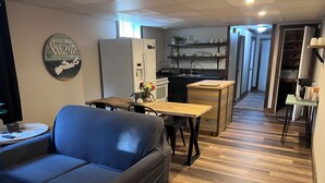 Ein Wohnzimmer mit einer Couch, einem Esstisch und einer Küchenzeile inklusive Kühlschrank und Regalen.