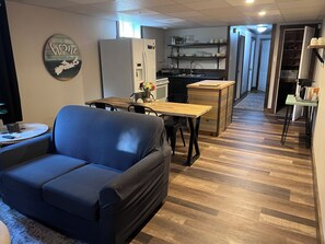 A living room with a sofa, a dining table, and a kitchen area with a refrigerator and shelves. - Charmer Down Under (Kentville)