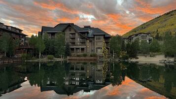 Un hôtel à plusieurs étages dont l’image se reflète dans un lac paisible, entouré d’arbres et d’un paysage vallonné.