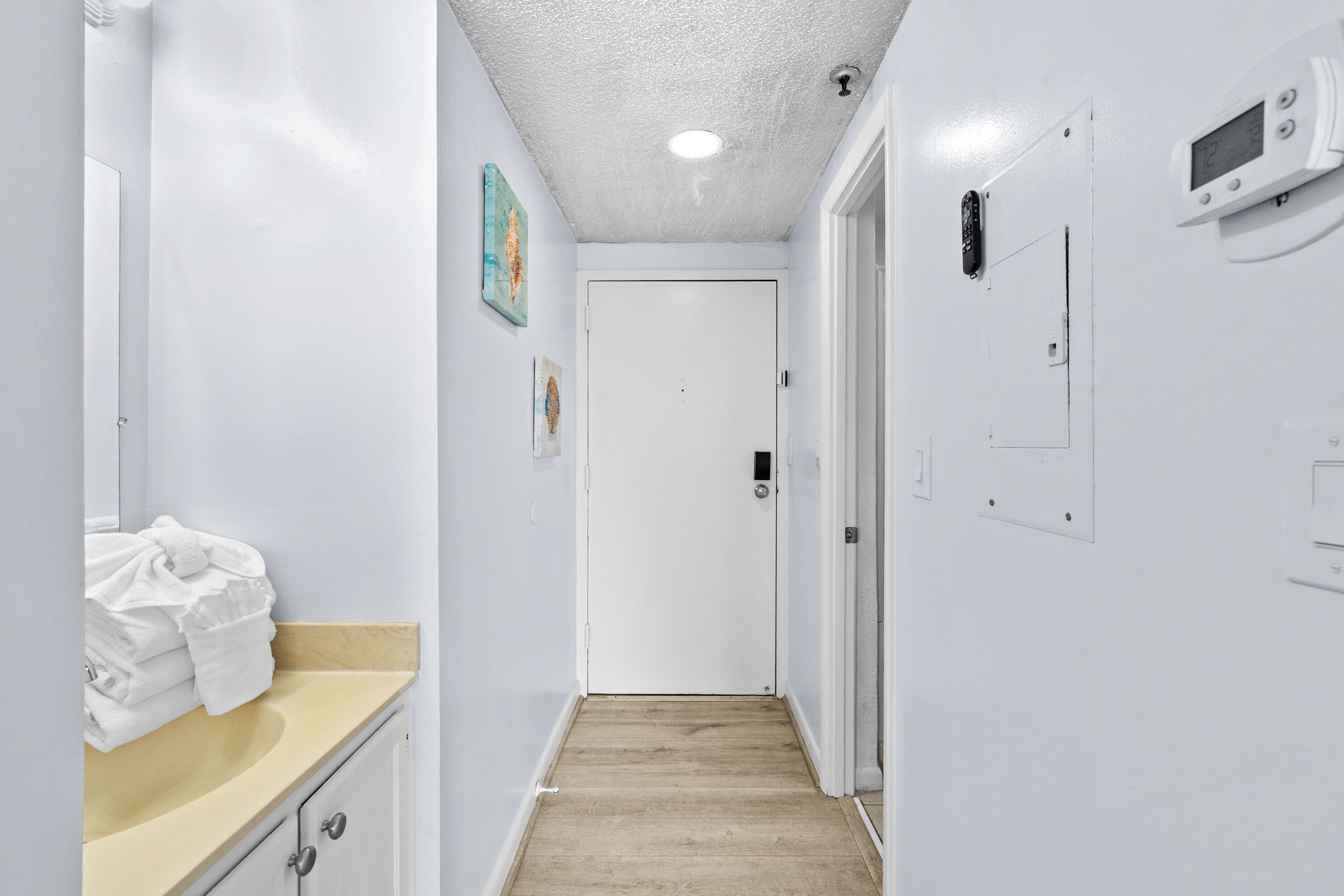 Un pasillo estrecho con suelo de madera, paredes blancas, un baño con lavamanos y toallas, y una puerta al final.
