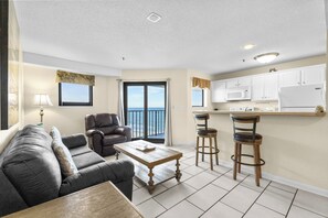 A living room with a sofa, coffee table, and two chairs. There is a kitchen with white cabinets, a microwave, and a refrigerator. A window with curtains and a view of the ocean is visible. - Myrtle Beach Resort RT1213 (Myrtle Beach)