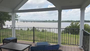 A covered outdoor area with a view of a bridge over water, furnished with wicker furniture and a table.
