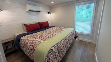 A bedroom with a bed, a window with blinds, an air conditioning unit, and a small bedside table.