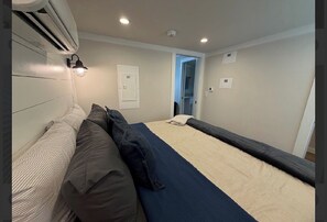 A room with a bed, a bedside table, an air conditioning unit, and a door leading to another room.