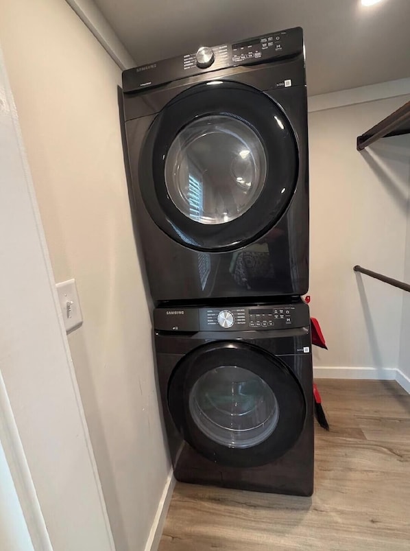 A stacked washer and dryer unit in a corner of a room with a wooden floor and white walls.