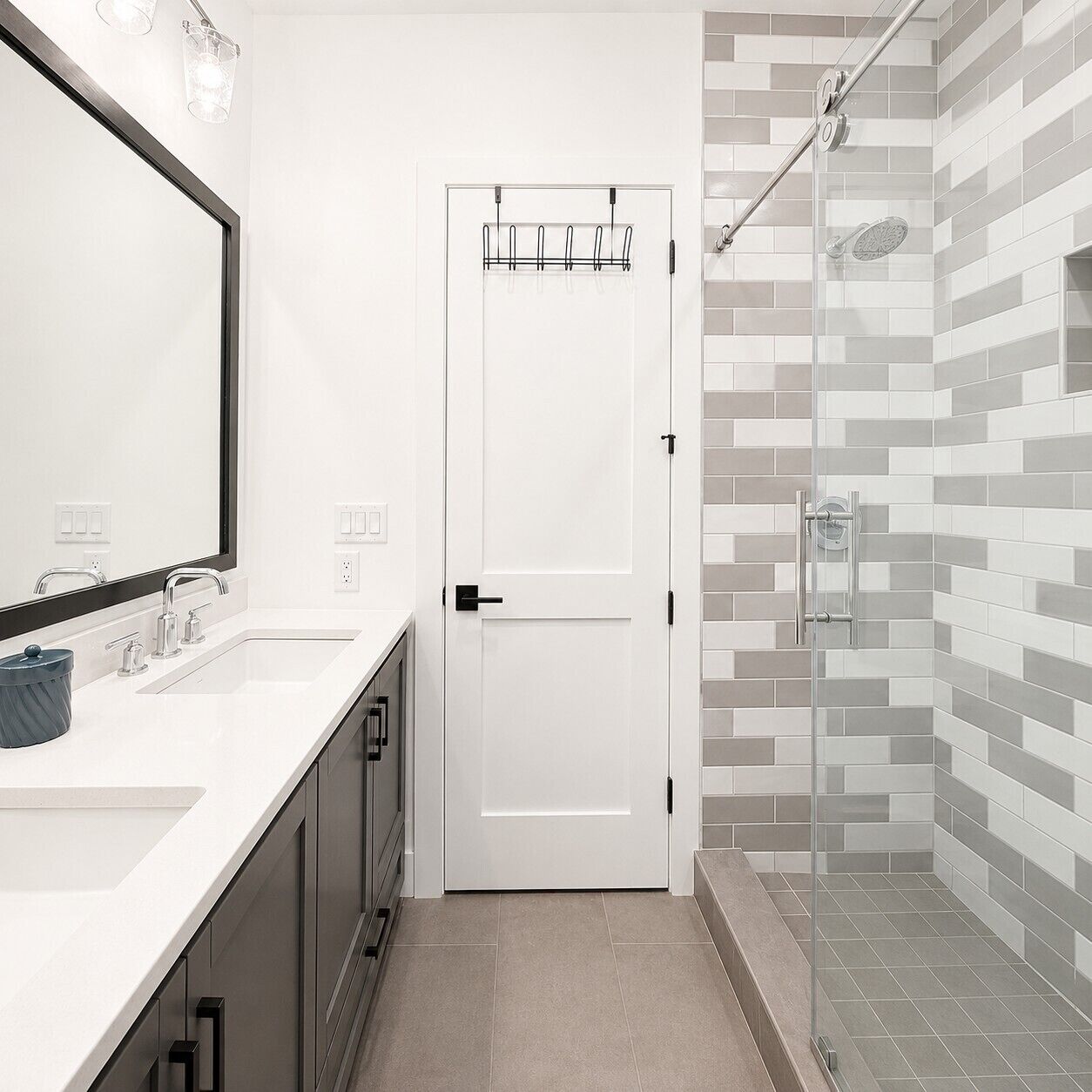 Une salle de bain avec un lavabo blanc, un grand miroir, une douche en verre et une porte dotée d’un porte-serviettes.