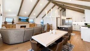 A modern kitchen with white cabinets, a dining area with a light-colored table and brown chairs, and a living area with a gray sofa and a fireplace. - Firework Views from the Private Hot Tub, Luxury Sierra Colina Retreat (SC109) (Stateline)