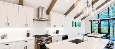 Una cocina moderna con armarios blancos, electrodomésticos de acero inoxidable, una isla grande con asientos y vistas a árboles a través de ventanales.