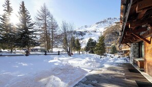 A snow-covered wooden deck leading to a building with a stone facade, surrounded by trees and a mountain in the background.