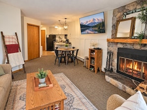 A cozy living room with a fireplace, a dining area, and a kitchen in the background.