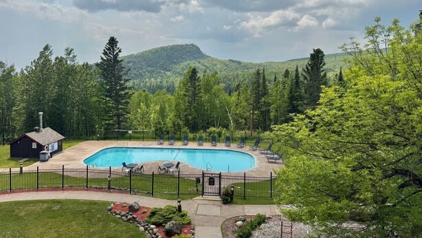 A swimming pool with lounge chairs, a small building, and a forested mountain backdrop. - Aspen Cottage Ski In Ski Out 1BR at Lutsen Mtn (Lutsen)