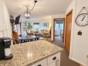 A kitchen with a coffee maker, a dining area with a table and chairs, and a living room with a sofa and a TV.