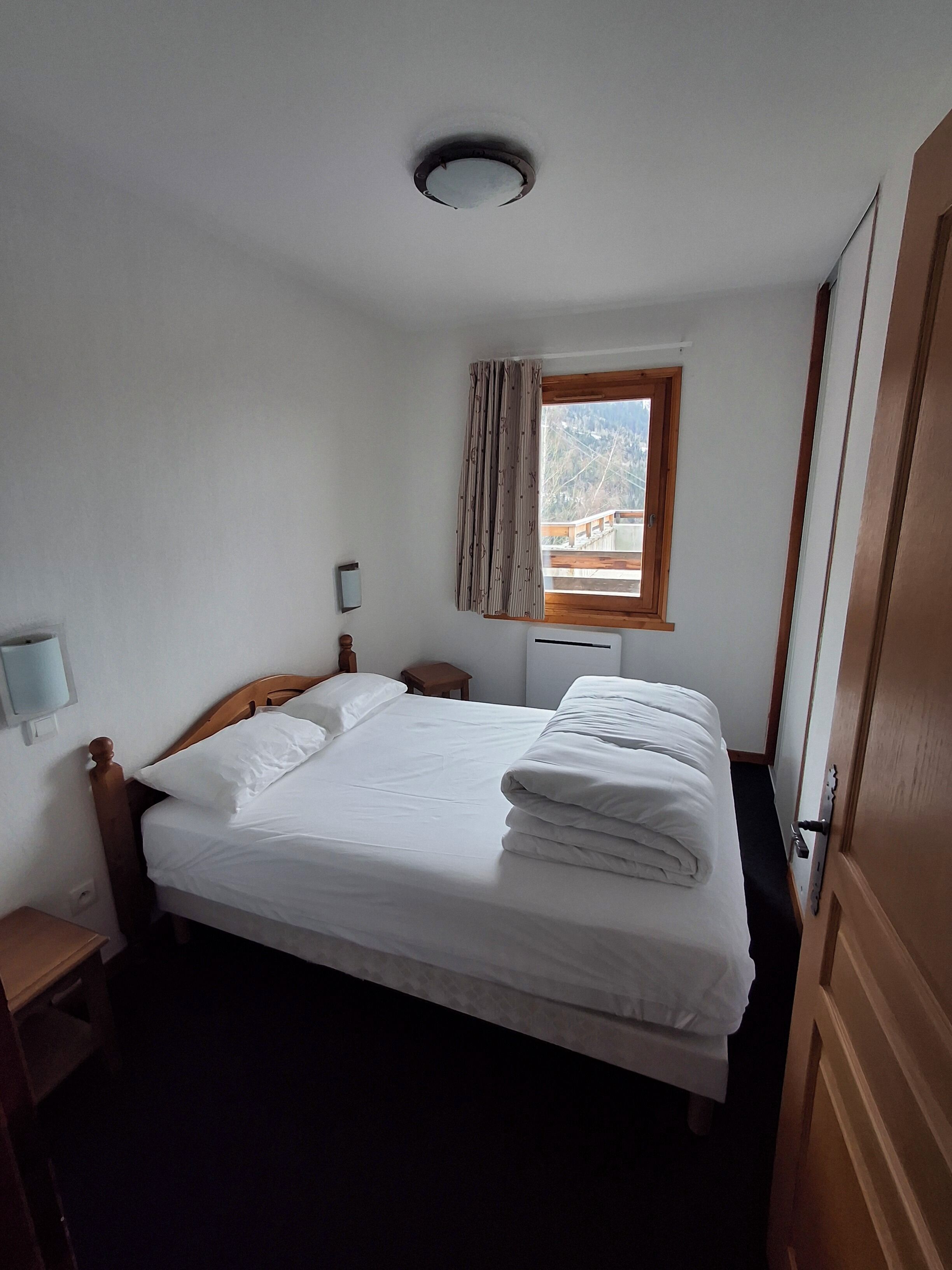 A hotel room with a bed, a window with curtains, a bedside table, and a wall-mounted light.