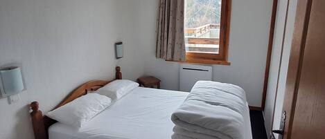 A hotel room with a bed, a window with curtains, a bedside table, and a wall-mounted light.