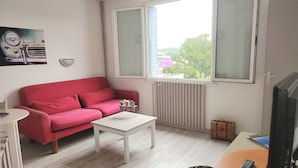 Una habitación con sofá rojo, una mesa de centro blanca, televisor y ventana con vista a árboles y edificios.
