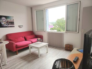 Ein Zimmer mit einem roten Sofa, einem weißen Couchtisch, einem Fernseher und einem Fenster mit Blick auf Bäume und Gebäude.