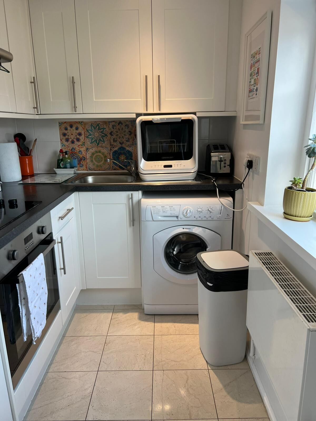 A compact kitchen with a built-in oven, a microwave, a washing machine, and a trash bin.