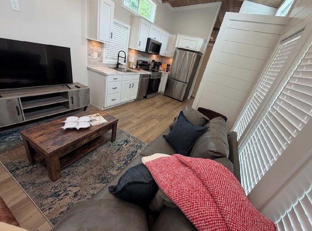 Uma sala de estar moderna com televisão de tela plana, mesa de centro de madeira, sofá com manta vermelha e área de cozinha com armários brancos e eletrodomésticos de aço inoxidável.