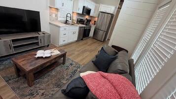 Un salon moderne avec un téléviseur à écran plat, une table basse en bois, un canapé recouvert d’un plaid rouge et un coin cuisine doté de meubles blancs et d’appareils électroménagers en acier inoxydable.