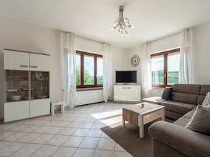 Ein modernes Wohnzimmer mit Sofa, Fernseher, Essbereich und großen Fenstern.