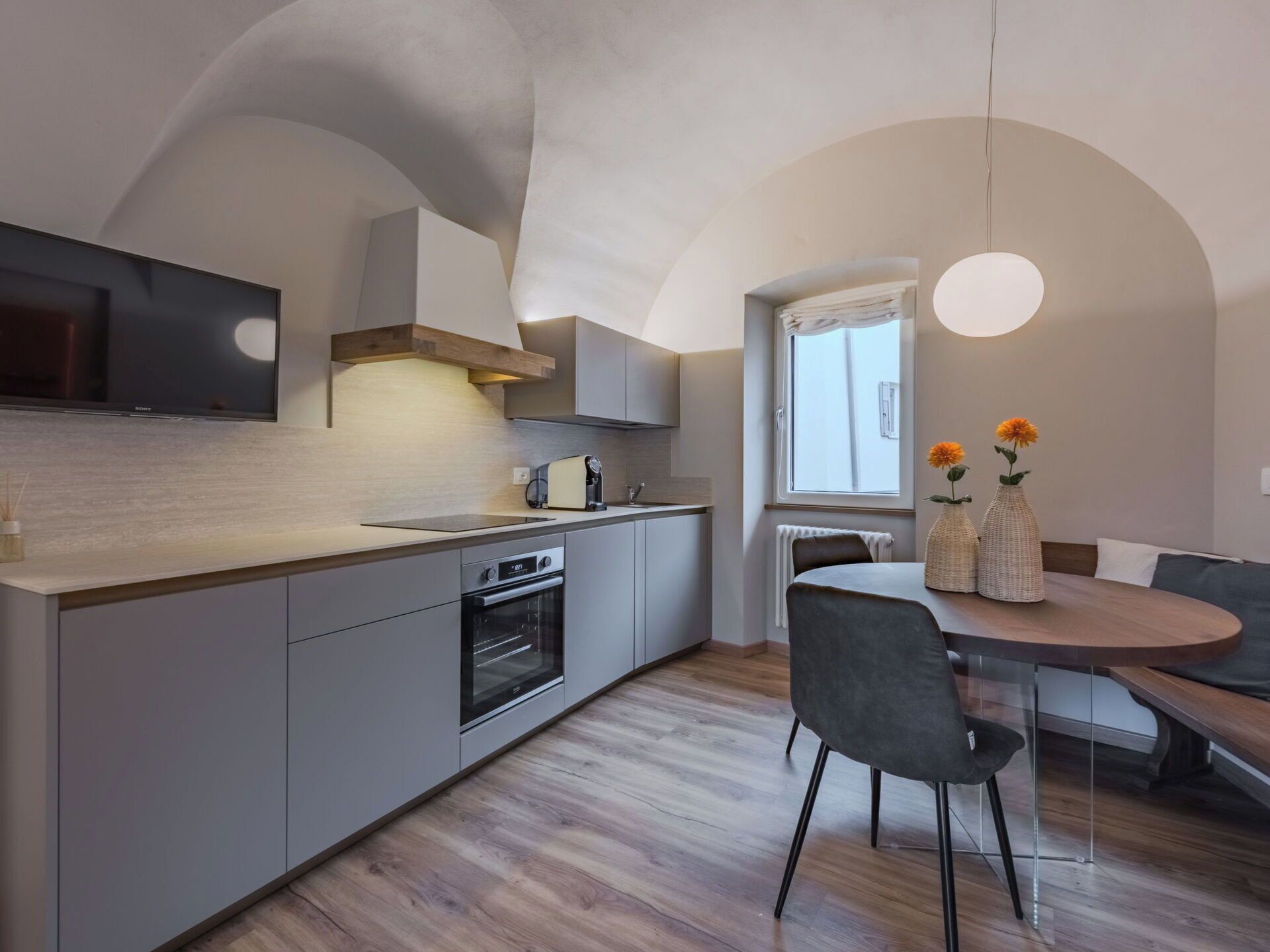 A modern kitchen with a built-in oven, a dining area with a round table and chairs, and a large window with curtains.