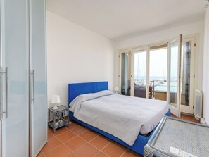 Un dormitorio con cama, mesita de noche, lámpara, vista al mar a través de una puerta corredera y balcón con mesa.