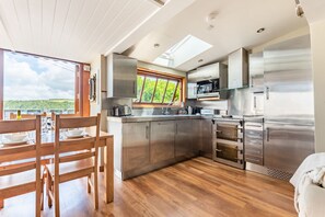 A modern kitchen with stainless steel appliances, a dining area with wooden furniture, and a large window offering a view of greenery. - Castle House Studio - Three Bedroom Cottage, Sleeps 6 (Fowey)