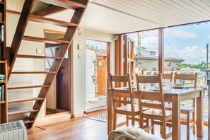 A wooden staircase leading to a door, a dining area with wooden chairs and a table, and a view of a stone building through large windows. - Castle House Studio - Three Bedroom Cottage, Sleeps 6 (Fowey)