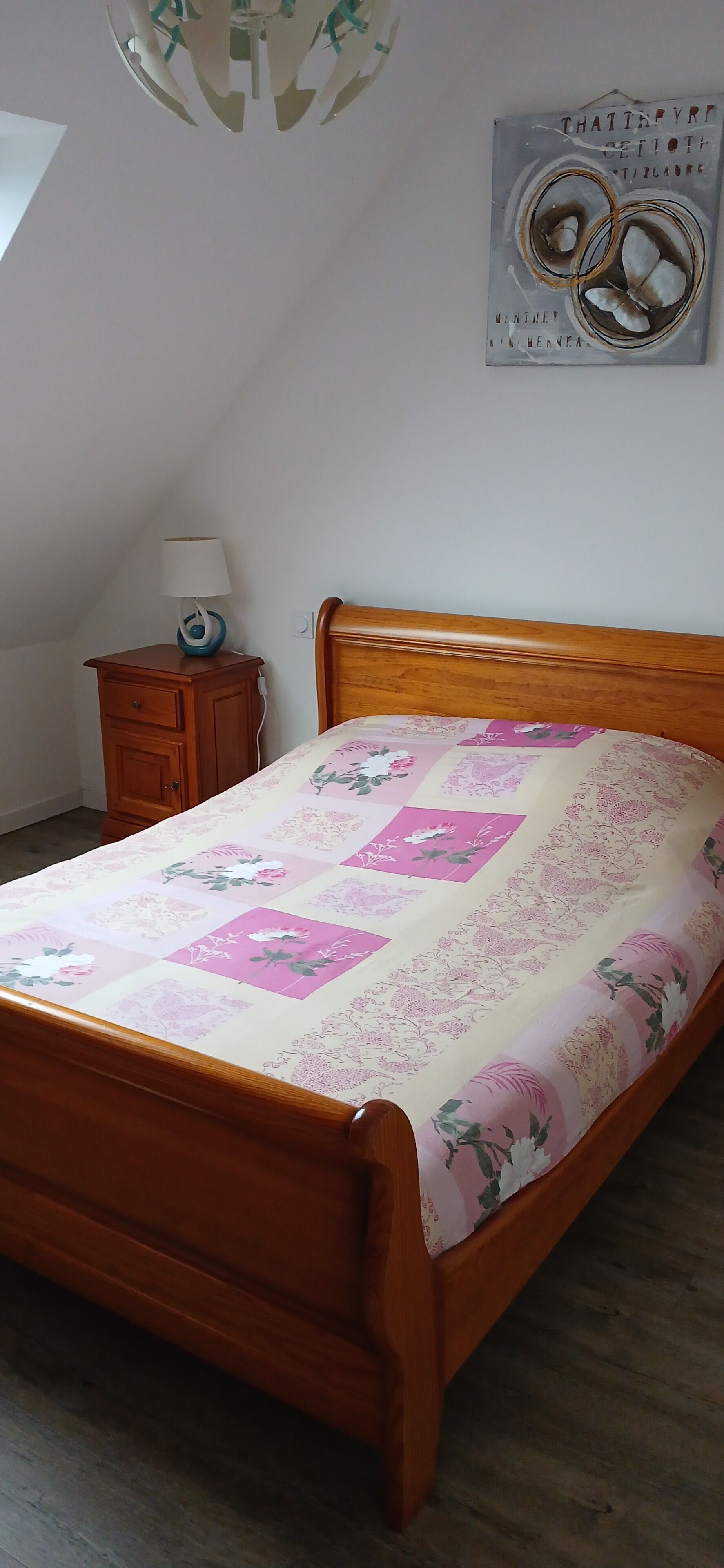 A bedroom with a wooden bed, a nightstand with a lamp, a wall clock, and a ceiling light fixture.