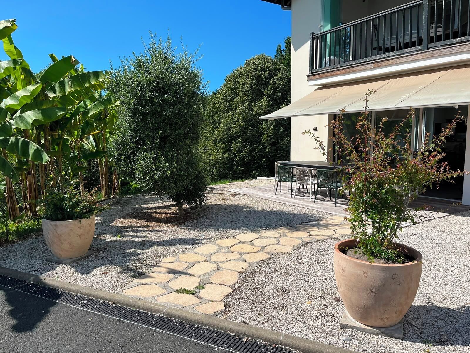 Une terrasse dallée avec une table et des chaises, entourée de plantes en pot et bordée d’un bâtiment doté d’un balcon.