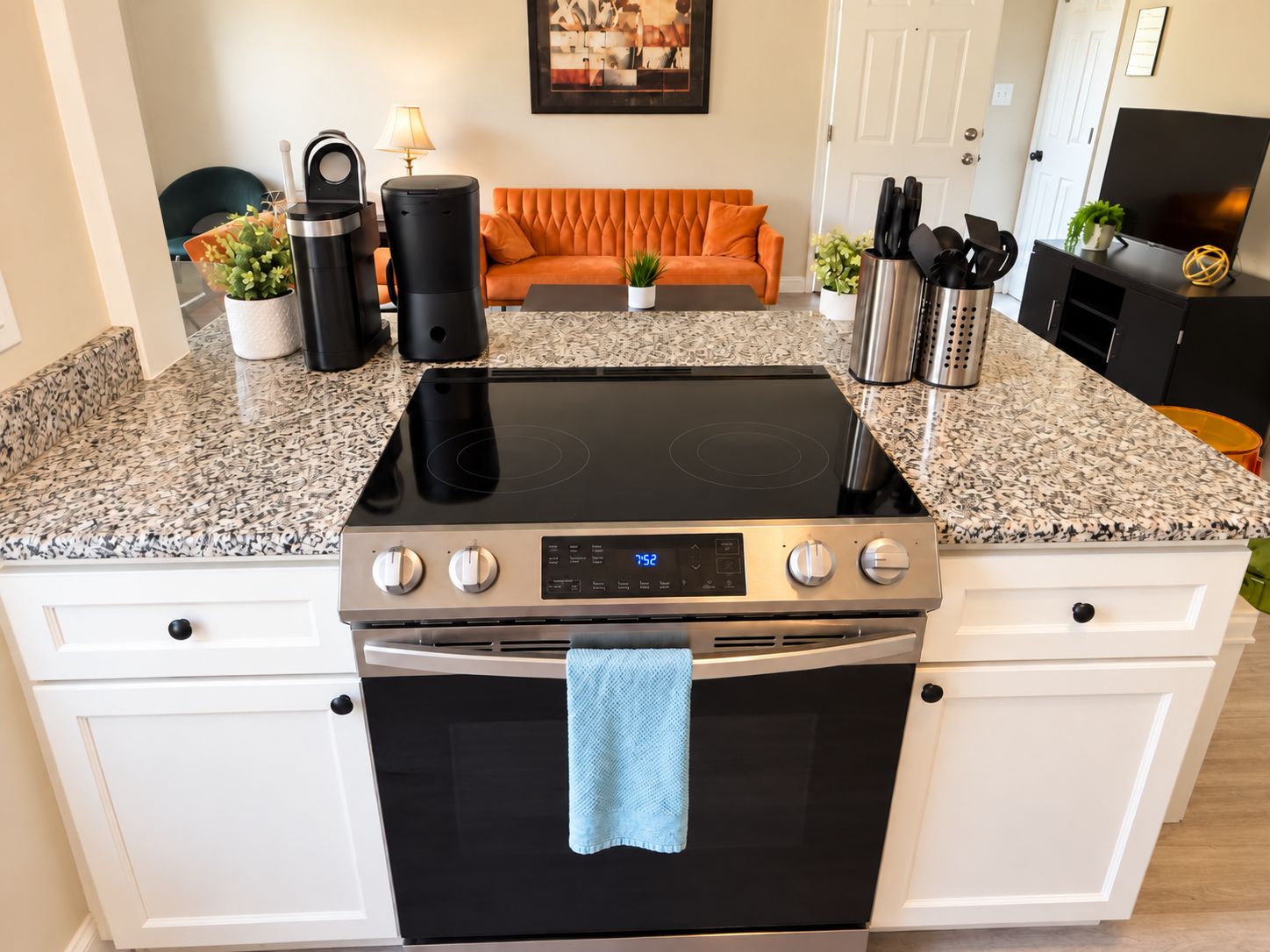 A kitchen with a black induction cooktop, a stainless steel oven, white cabinetry, a blue towel hanging on the oven door, and a countertop with a coffee maker, kettle, and other appliances.