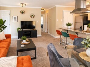 A living room with a sofa, coffee table, and TV. There is a kitchen area with a stove, sink, and bar stools. - The Durham Skybox (Durham)