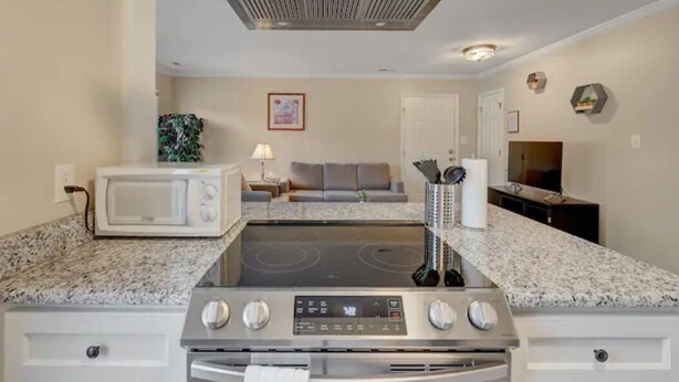 Una cocina moderna con microondas, estufa y horno integrados. Se observa un sofá gris y una pequeña mesa con una lámpara al fondo.