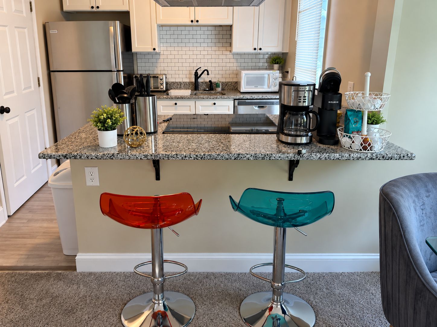 A kitchen with a stainless steel refrigerator, a microwave, a coffee maker, and a kettle. There are two stools with red and teal seats.