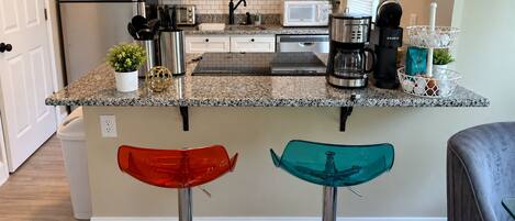 A kitchen with a stainless steel refrigerator, a microwave, a coffee maker, and a kettle. There are two stools with red and teal seats.