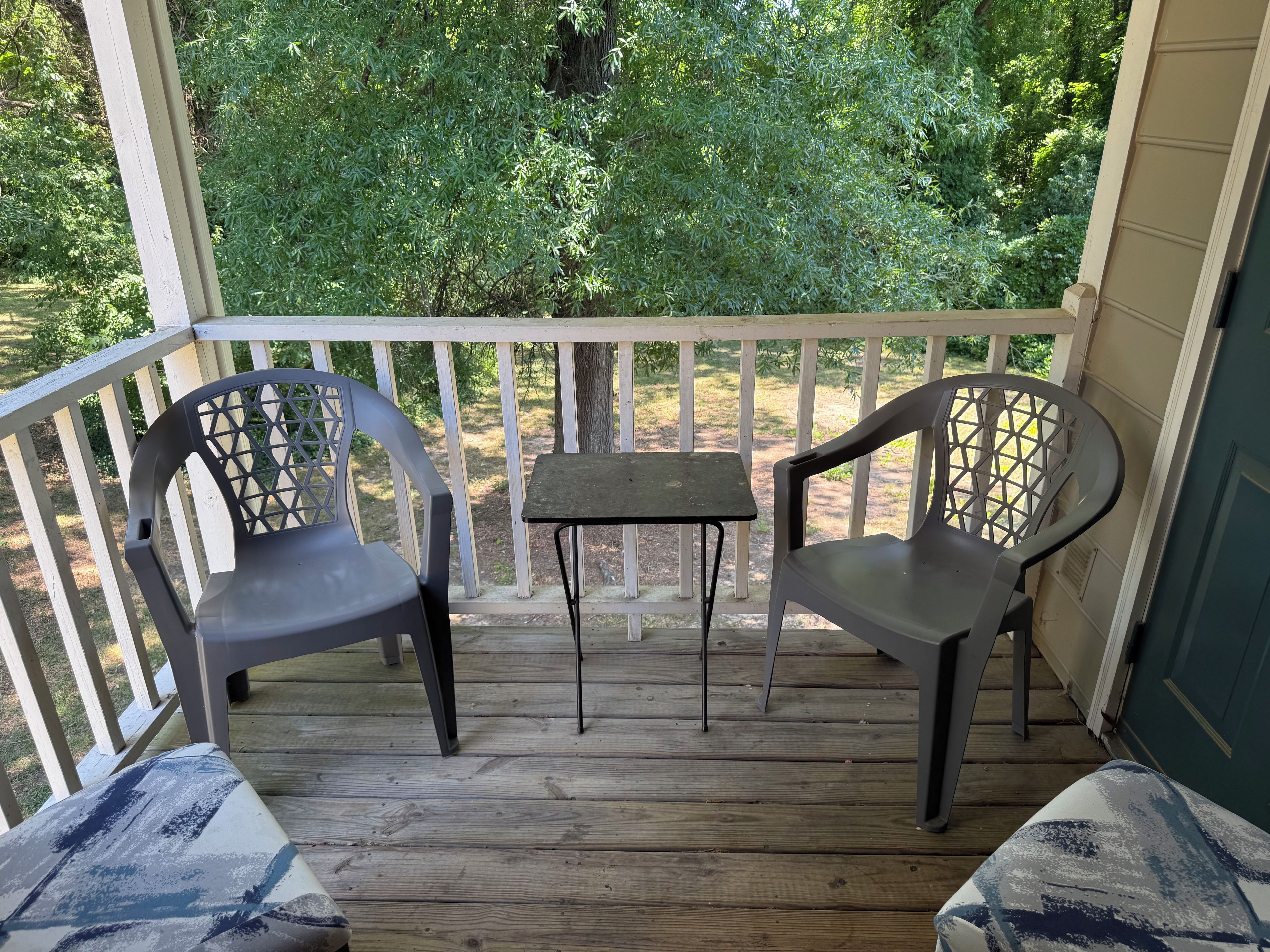 A wooden deck with two chairs and a small table, surrounded by a white railing, overlooking a green landscape.
