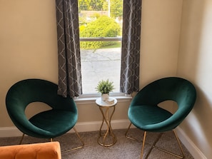 A room with two teal chairs, a small white table with a plant, and a view of a green landscape through a window with patterned curtains. - The Durham Skybox (Durham)