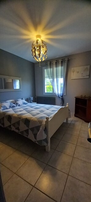 A bedroom with a bed, a chandelier, a window with curtains, a radiator, and a small cabinet.