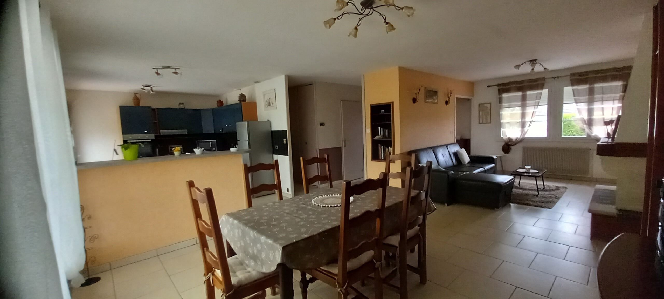 Ein Wohnzimmer mit einer Couch, einem Esstisch mit Stühlen, einer Küchenzeile inklusive Schränken und Geräten sowie einem Fenster mit Vorhängen.