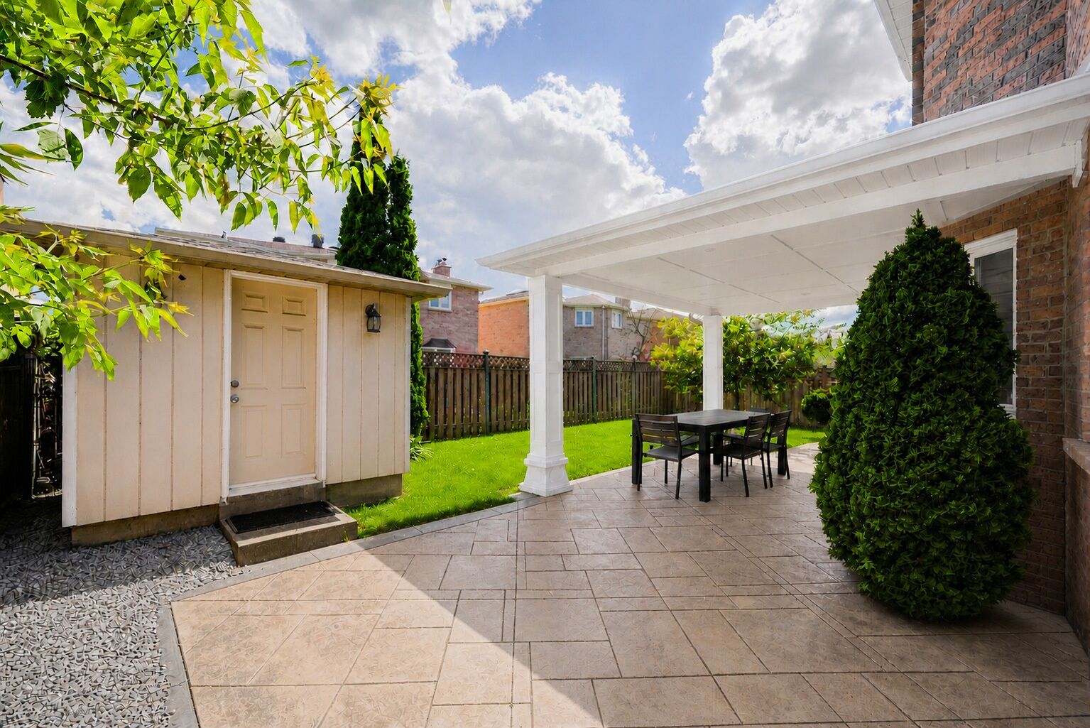 Ein überdachter Patio-Bereich mit Tisch und Stühlen, ein kleines Gartenhaus und eine gepflegte Rasenfläche.