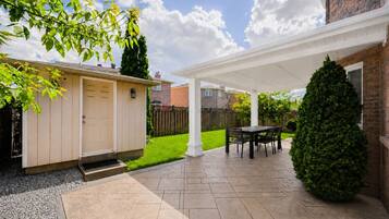Ein überdachter Patio-Bereich mit Tisch und Stühlen, ein kleines Gartenhaus und eine gepflegte Rasenfläche.