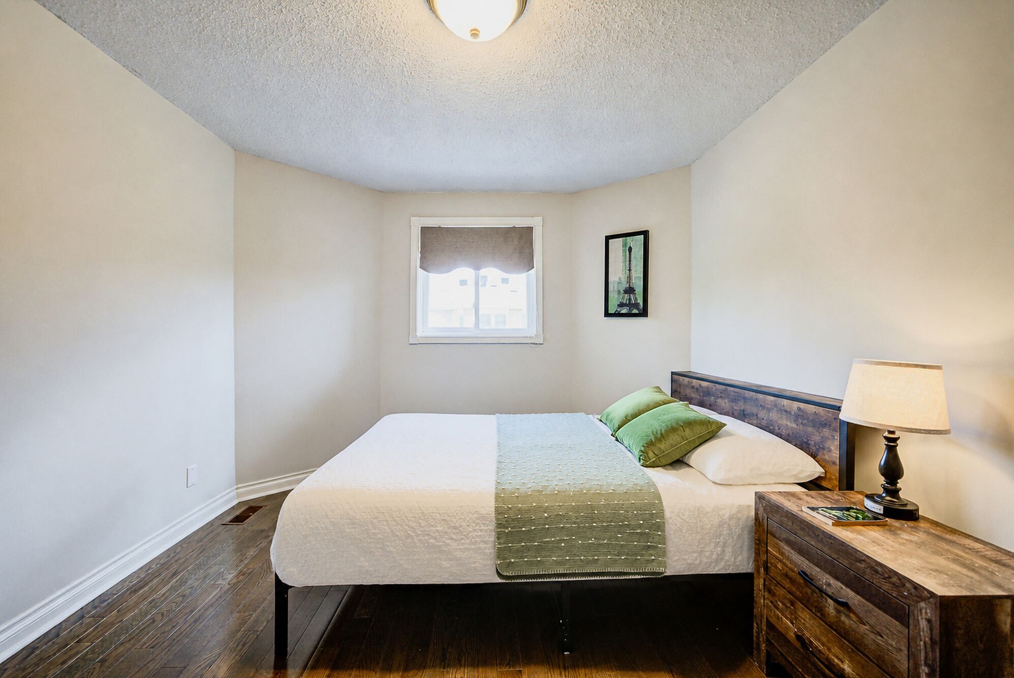 A bedroom with a bed, a nightstand, a lamp, a window, and a picture on the wall.